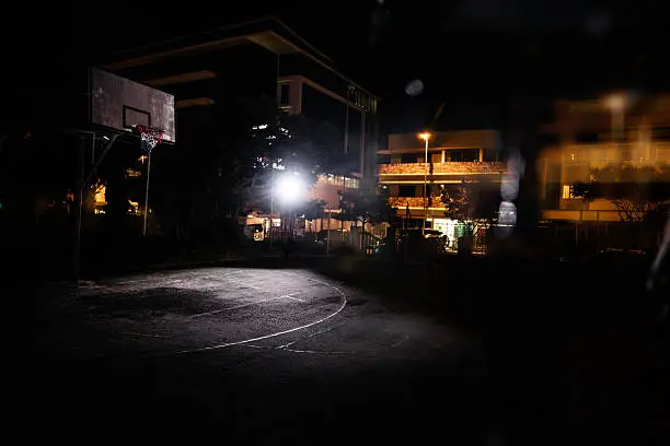 Establishing shot of empty inner-city basketball court illuminated by one direct light source and city lights in the background in the night