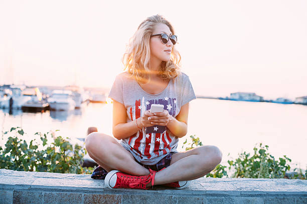 Young woman using smart phone on the beach Young blond woman using smart phone on the beach. Wear sunglasses, gray T-shirt with USA flag, shorts, and red sneackers. Sea and quay with sailboats as background. cross legged photos stock pictures, royalty-free photos & images