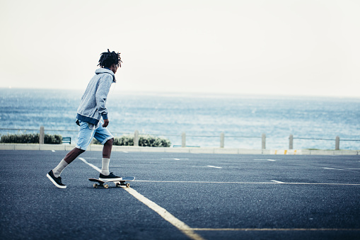 African American man in shorts and hoodie skating on his skateboard on an seaside parking lot with ocean in background