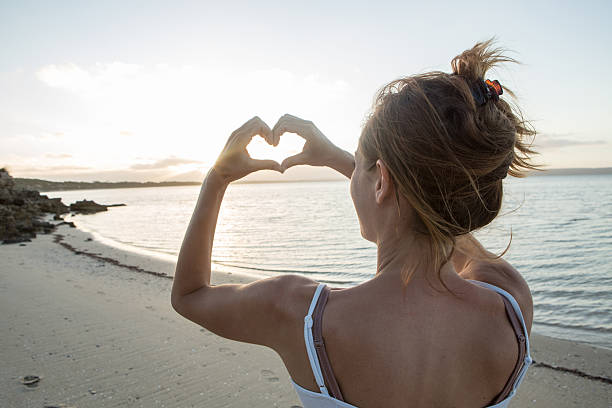 istock Woman on beach makes heart shape finger frame towards sun 518092596