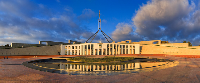 panorama of new Australian Parliament in Canberra, ACT. Facade of modern building reflecting in pond of fountain at sunrise