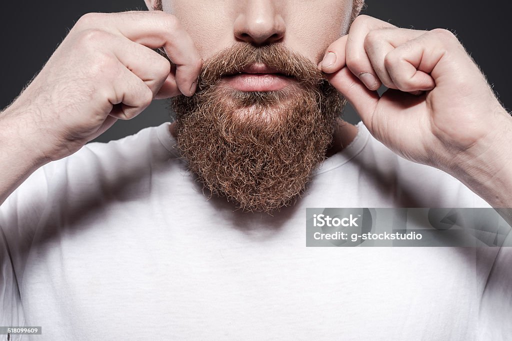 Making his own style. Close-up of young bearded man adjusting his mustaches while standing against grey background Adult Stock Photo