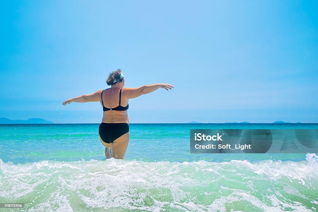 mature woman in swimwear at the sea. mature woman in swimwear at the sea. Overweight woman in swimsuit comes in water in time of surf, rear view Women Stock Photo