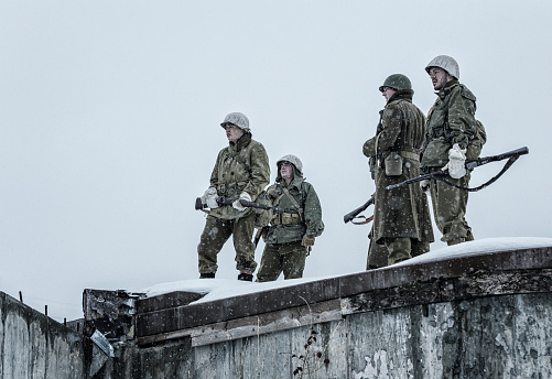 A platoon of WWII US Army combat infantry soldiers is standing on top of a burned out building concrete bunker perimeter wall during a winter snow blizzard. The men are looking curiously down at the damaged building as they wait for their next orders. Most of them wear a white cloth as makeshift winter camouflage covering their standard issue metal helmets, and the uniforms are wet from the heavy snowfall.