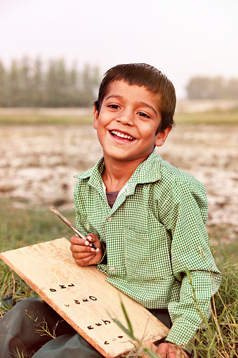 Little boy of Asian ethnicity sitting in the grass wearing school uniform & writing on takhti using Kalam & looking at camera portrait outdoor. In Old Time Primary School Students Bring Wood Made Board Called \