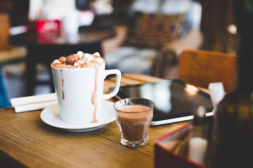 A cup of hot chocolate sits on a table next to a digital laptop. Topped with marshmallows and chocolate balls ready to be drank. A spoon lays on a napkin at the side. A shot glass with extra hot chocolate stands beside the cup as it overflows slightly.