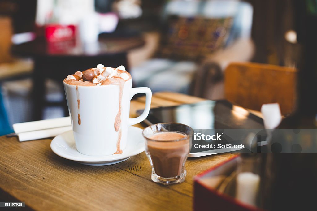 Luxury hot chocolate for one! A cup of hot chocolate sits on a table next to a digital laptop. Topped with marshmallows and chocolate balls ready to be drank. A spoon lays on a napkin at the side. A shot glass with extra hot chocolate stands beside the cup as it overflows slightly. Hot Chocolate Stock Photo
