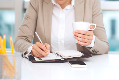 Business woman writing in a schedule planner, and drinking coffe