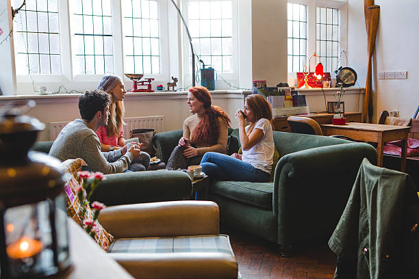 Friends gather around a low table together in a coffee shop. One girl holds a coffee cup, food is on the table and faces of two can be seen. Sideview of their friends. Large windows in the background letting in light. Lower angle view and other chairs can be seen