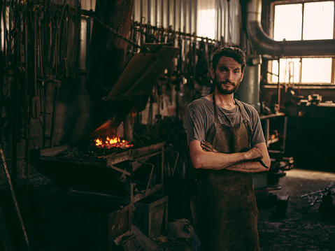 Blacksmith portrait. Young adult bearded man standing in his workshop showing fullfilment with folded arms.