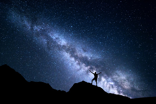 Landscape with Milky Way. Night sky with stars and silhouette of a happy man with backpack and raised up arms on the mountain. Space background