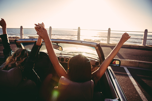 Two happy young girl friends celebrating with raised arms in a convertible parked in front of the beach at sunset