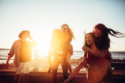 Fun-loving friends having piggyback rides on a promenade in summertime with sunset sun flare