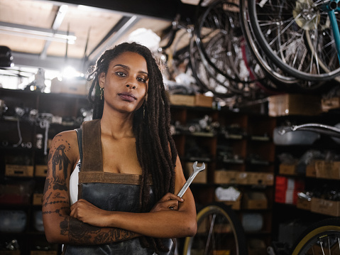 Portrait of an afro-american woman who is a bicycle mechanic with tattoos and dreadlocks standing with her arms crossed and looking proudly at the camera in her bike repair workshop with bicycles, tools and parts all around her