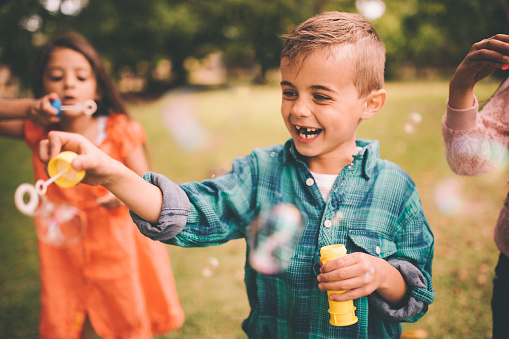 Cute little boy laughing and playing by making bubbles with a bubble wand in a summer park with friends