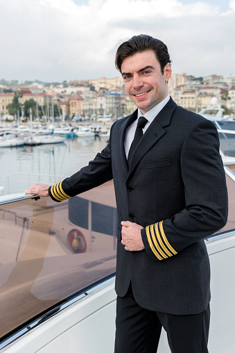 Vertical color image of young smiling boat captain standing on luxury yacht top, wearing elegant uniform and looking at camera. In background sea and port of Cannes.