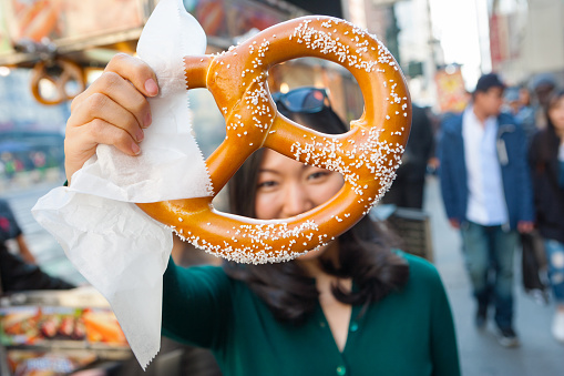 Young attractive Asian girl holding a pretzelup to the camera, she purchased from street vendor food cart in NYC.