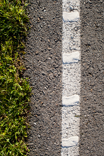 Close up of roadside, painted single white line on asphalt and green grass in the sunlight, view from above.