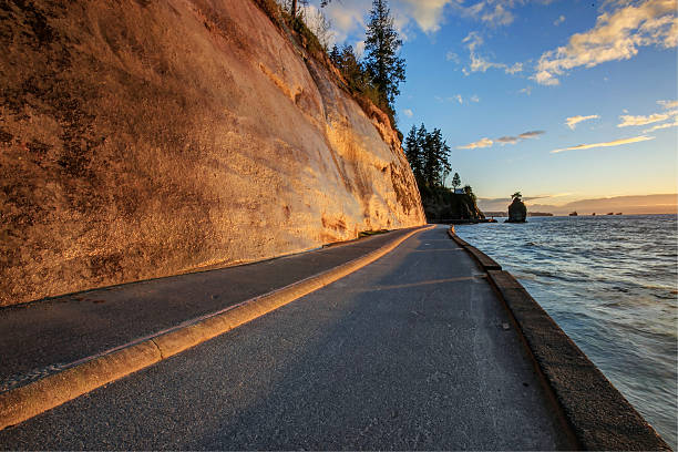 Calm waterfront pathway in Stanley Park with rocky shore and cargo ships visible.