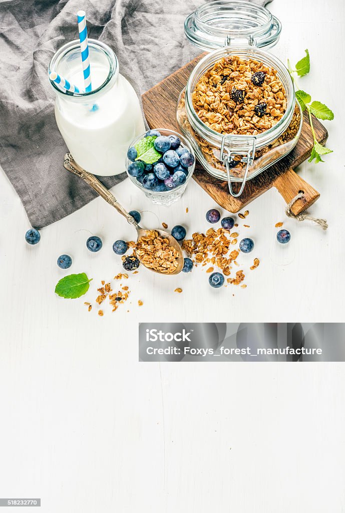 Healthy breakfast ingrediens. Homemade granola in open glass jar, milk Healthy breakfast ingrediens. Homemade granola in open glass jar, milk or yogurt bottle, blueberries and mint on white wooden background, top view, copy space, vertical Breakfast Stock Photo