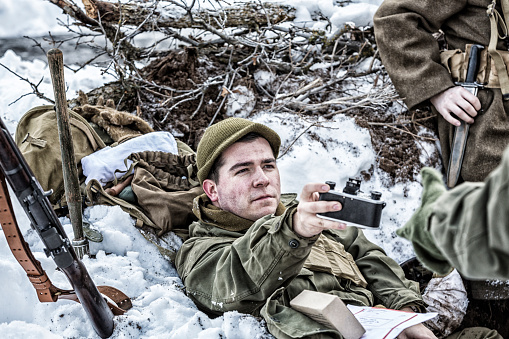 Relaxing in a frozen foxhole in a winter snow covered bunker, a World War II US Army combat infantry soldier is passing his authentic 1940s era film camera to his buddy for assistance with some snapshots of himself and his platoon comrades.