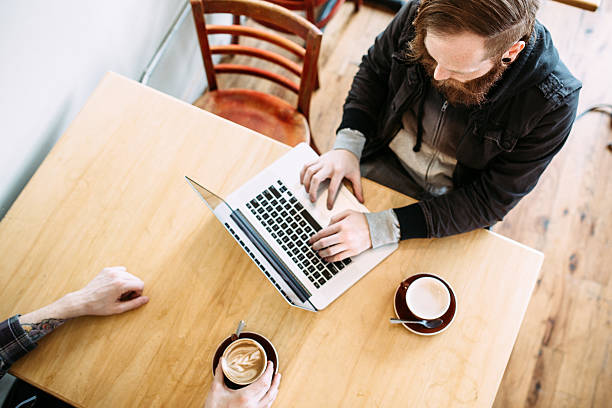 A young adult man in hipster style and fashion enjoys some relaxation in a nice urban cafe, drinking a single origin espresso while working on his laptop.  His friend sits across from him.  Shot from high above looking down.