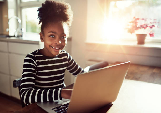 Smiling friendly young black african girl Smiling friendly young black african girl with a cute top knot afro hairstyle sitting at a laptop computer at the dining table grinning at the camera topknot stock pictures, royalty-free photos & images