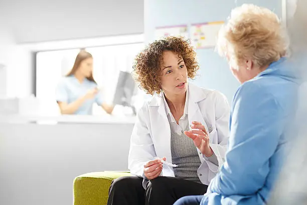 A female pharmacist sits with a senior female patient in the pharmacist consultation area and discusses her prescription and choice of medication. In the background a father and daughter stand at the dispensing counter and are served by a female pharmacy assistant .