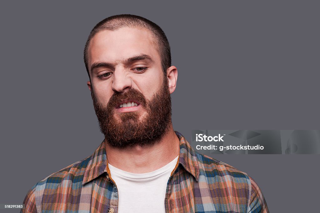 In friends with irony. Handsome young bearded man keeping arms crossed and expressing ironic smile while standing against grey background Disgust Stock Photo