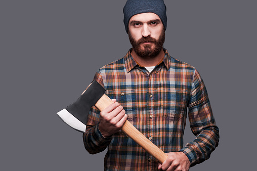 Confident young bearded man holding a big axe and looking at camera while standing against grey background