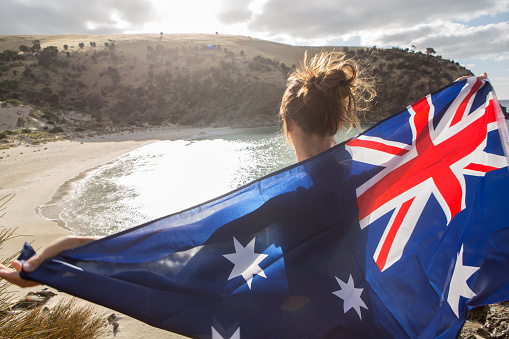 Cheerful young woman stands on a cliff above a beach in Kangaroo Island holding an Australian's flag.