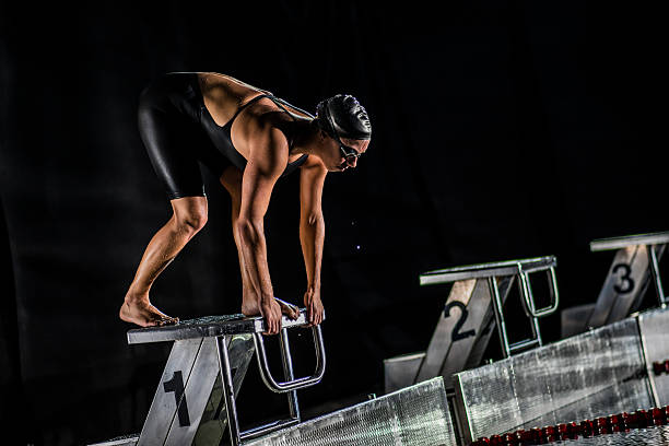 Swimmer On A Swimming Starting Block Professional female swimmer standing on the starting block and preparing to jump into an olympic pool.   start block stock pictures, royalty-free photos & images