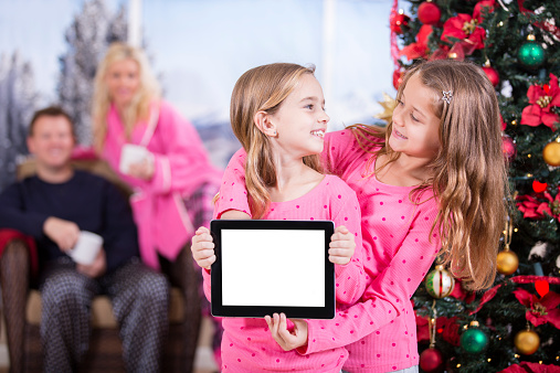 Cute little girl excitedly opens a digital tablet present on Christmas morning. Christmas tree, family in background.  White, blank digital display screen for copyspace. 