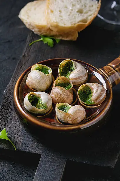 Escargots de Bourgogne - Snails with herbs butter, gourmet dish, in traditional ceramic pan with parsley and bread on wooden chopping board over black textured background.