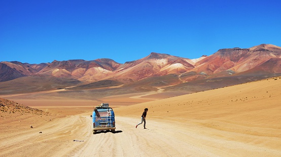 Uyuni, Bolivia - March 28, 2016: A blue classic Vw Bus parked on a sandy path in front of colorful mountains - A woman walking away from the bus