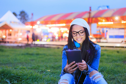 Young girl looking at digital tablet and blue headphones in amusement park by night. Wear denim jacket, white cap and black eyeglasses.
