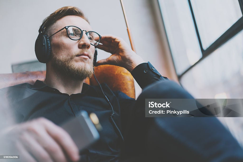 Portrait handsome bearded man glasses,headphones listening to music modern Closeup photo handsome bearded man glasses in headphones listening to music modern loft studio.Man sitting in vintage chair, holding smartphone and relaxing.Panoramic windows background,film effect.  Lifestyles Stock Photo