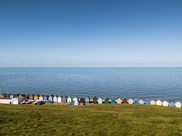 Line, row of beach huts Herne bay, Kent, uk A long row, line of small wooden beach huts in Herne Bay, Kent, at the bottom of a green grass slope in front of blue sea with wind turbines on the horizon. herne bay photos stock pictures, royalty-free photos & images