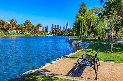 A lake with rippling waters and a park bench fill the foreground leading back to the skyscrapers of downtown skyline Los Angeles, California