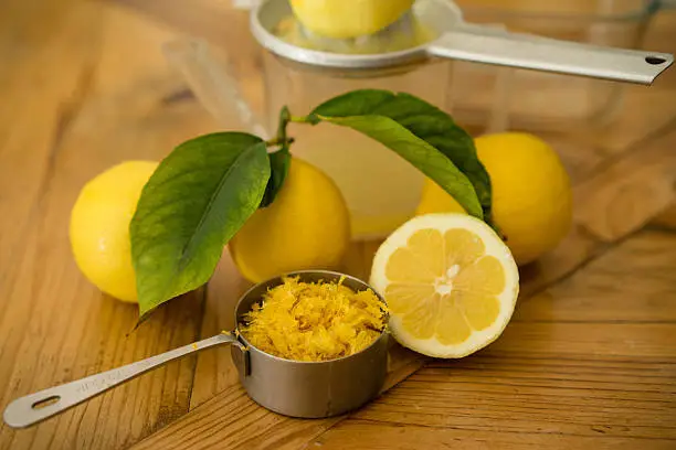 Fresh lemon zest in a bowl next to a sliced lemon and two whole lemons with leaves attached sitting on a wooden background.