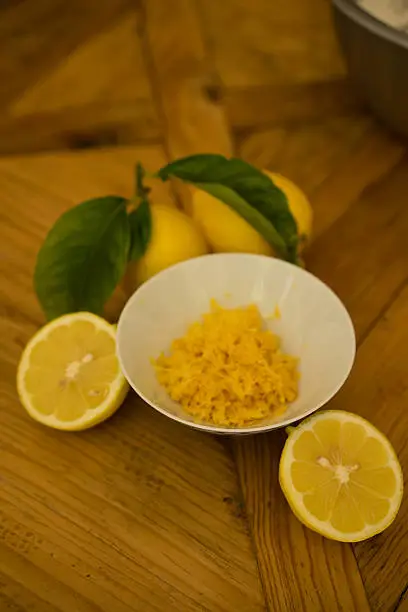 Shallow focus of fresh lemon zest in a white bowl, cut lemon and two lemons on the stem with a wooden background.