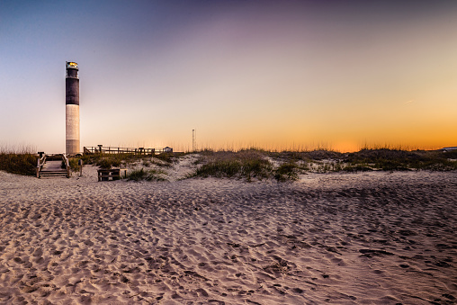 This is an image of the monumental lighthouse of Oak Island, in Brunswick County, North Carolina, taken early in the morning during spring season. The area consists of beautiful beaches for swimming and several wharfs for fishing. This shot was taken viewing from the beach, with the beautiful texture of the white sand in the foreground. Oak Island is just a few miles away from Wilmington, the home of NBA legend Michael Jordan.