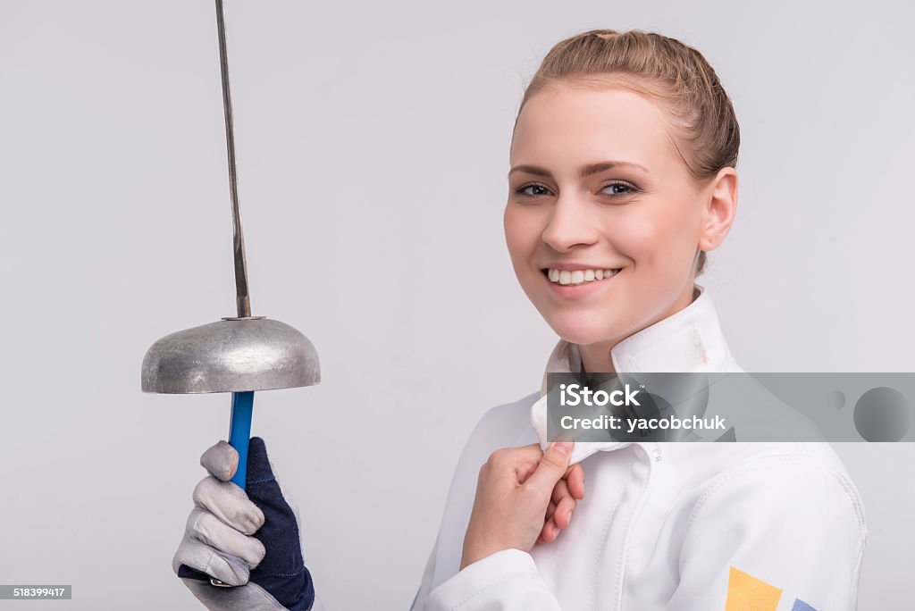 Young woman engaging in fencing Half-length portrait of happy young smiling woman wearing white fencing costume holding sword in her hand standing aside satisfied with her win. Isolated on white background Adult Stock Photo