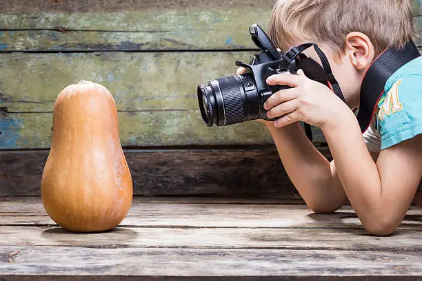 Funny fair boy use DSLR camera trying to take photo of ripe pumpkin on wooden background in studio. Photography lessons or learning background