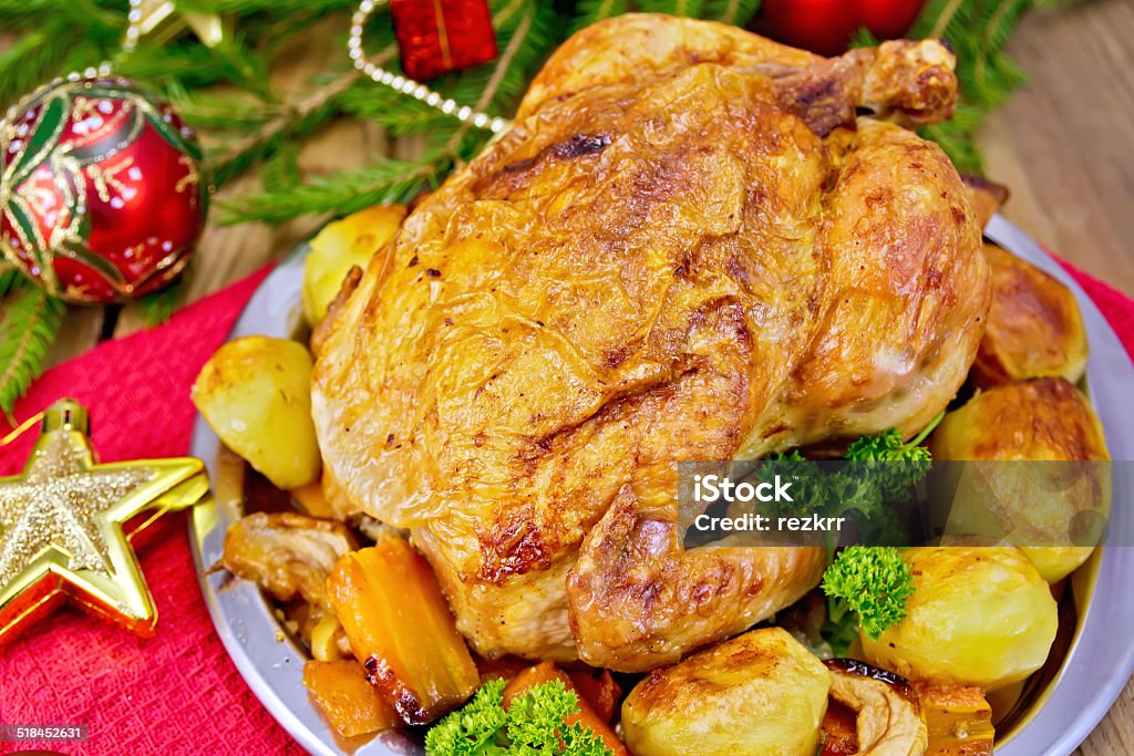 Chicken with red Christmas toys on board Chicken baked with vegetables and apples on a metal plate, red and gold Christmas toys, napkin, spruce twigs on a wooden boards background Apple - Fruit Stock Photo