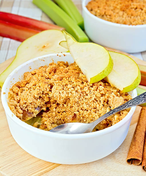 Crumble with pears and rhubarb in a white bowl on a wooden board, rhubarb stalks and pear, cinnamon on a background of striped linen tablecloth