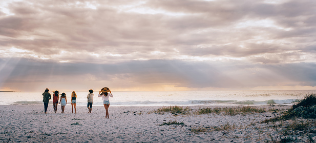 A Hipster multi-ethnic group of friends walking barefoot and happy on sandy beach towards sun beam lit ocean
