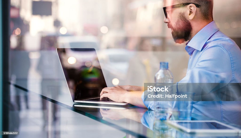 Hipster manager in cafe, working on laptop by window Hipster manager sitting in cafe by the window working on laptop, street reflection Blue Stock Photo