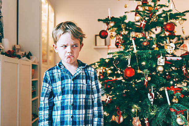 Unhappy boy stands before chistmas tree and makes a face Boy wears pajamas and stands before a decorated christmas tree on christmas morning. He looks unhappy and makes a face. disappointment photos stock pictures, royalty-free photos & images