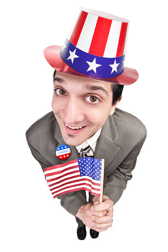 A fisheye image of a patriotic American man holding a flag and wearing a stars and stripes hat and a pin that reads \